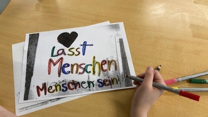 Eine Hand schreibt mit bunten Farben "Lasst Menschen Menschen sein".