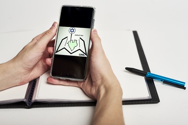 EIne Mappe und ein Stift. Davor Hände mit einem Handy. Das Handy zeigt das Logo von "Jüdisch hier". | LWL-Medienzentrum, Phillina Zuther
