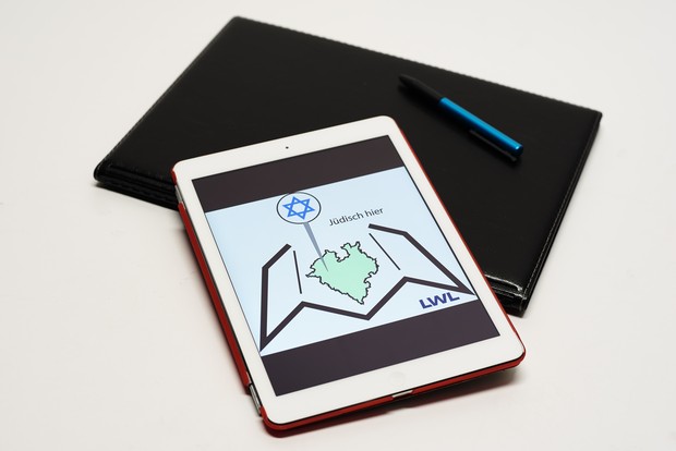 Ein Tablet, eine Mappe, ein Stift. Das Tablet zeigt das Logo von "Jüdisch hier" | LWL-Medienzentrum, Phillina Zuther Ein Tablet, eine Mappe, ein Stift. Das Tablet zeigt das Logo von "Jüdisch hier" | LWL-Medienzentrum, Phillina Zuther